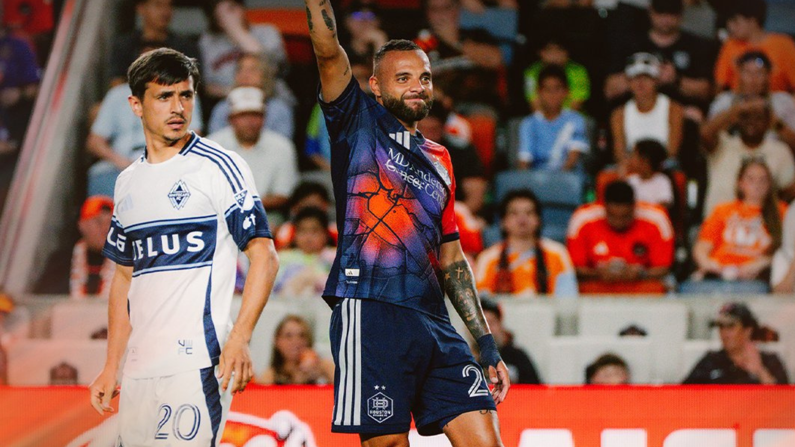 Two soccer players on the field during a match; one in a white jersey with number 20, the other in a dark blue kit raising his arm amid a cheering crowd.