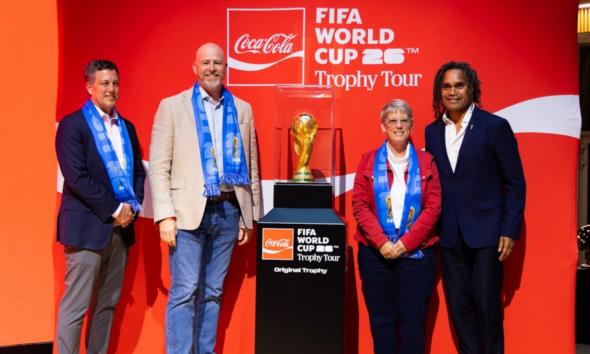 Four people stand on a red stage beside a pedestal displaying the FIFA World Cup Trophy, all wearing blue scarves, with Coca‑Cola and Trophy Tour branding in the background.