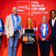 Four people stand on a red stage beside a pedestal displaying the FIFA World Cup Trophy, all wearing blue scarves, with Coca‑Cola and Trophy Tour branding in the background.