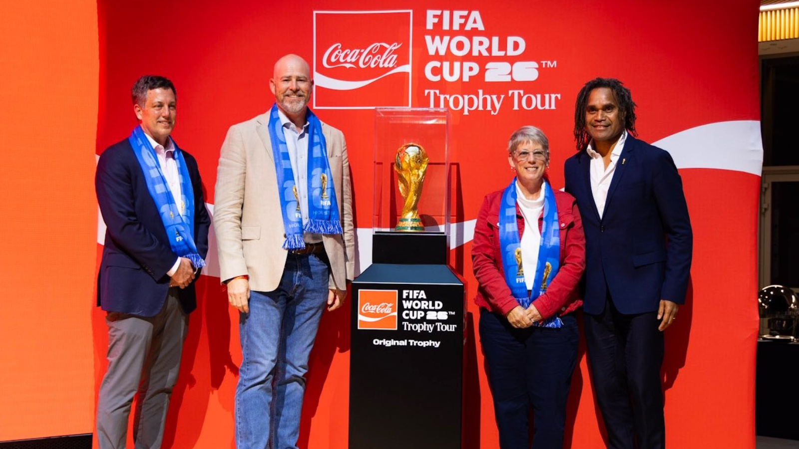 Four people stand on a red stage beside a pedestal displaying the FIFA World Cup Trophy, all wearing blue scarves, with Coca‑Cola and Trophy Tour branding in the background.