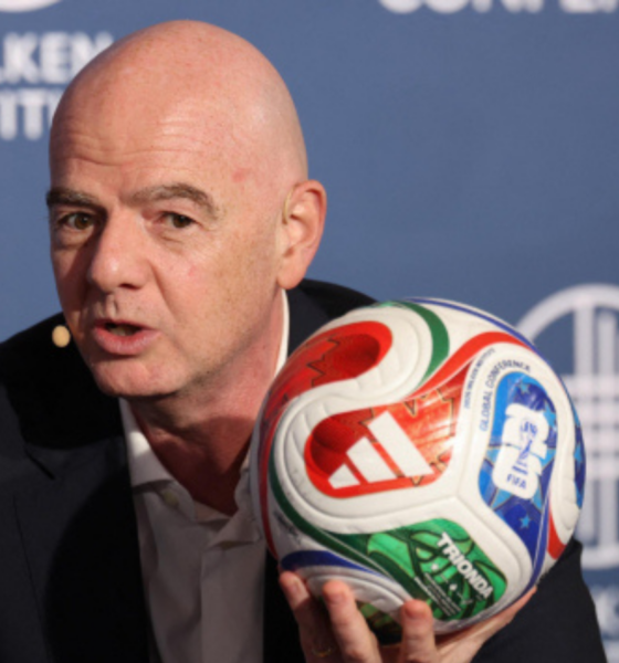 Bald man in a dark suit speaks at a Milken Institute event while holding a colorful soccer ball close to the microphone backdrop behind him.