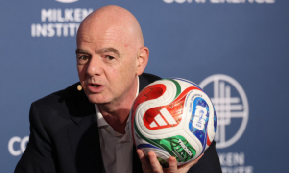 Bald man in a dark suit speaks at a Milken Institute event while holding a colorful soccer ball close to the microphone backdrop behind him.