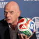 Bald man in a dark suit speaks at a Milken Institute event while holding a colorful soccer ball close to the microphone backdrop behind him.