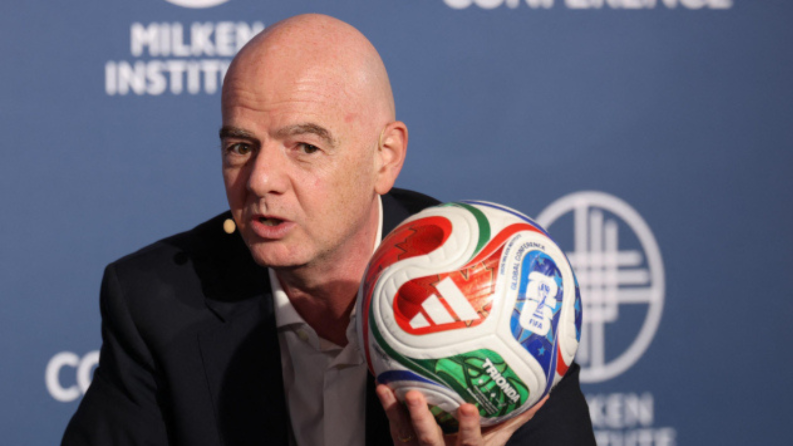 Bald man in a dark suit speaks at a Milken Institute event while holding a colorful soccer ball close to the microphone backdrop behind him.