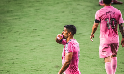 Two soccer players in pink uniforms on a green pitch; one touches a pink glove to his lips while the other walks away with the number 10 on his back.