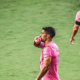 Two soccer players in pink uniforms on a green pitch; one touches a pink glove to his lips while the other walks away with the number 10 on his back.