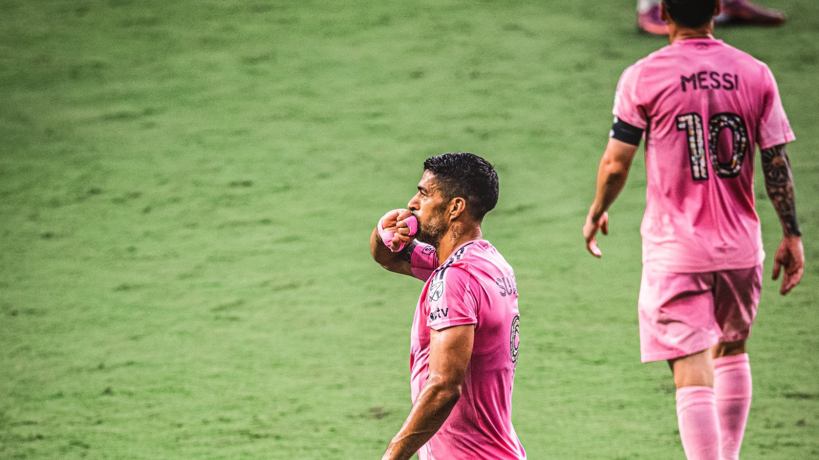 Two soccer players in pink uniforms on a green pitch; one touches a pink glove to his lips while the other walks away with the number 10 on his back.