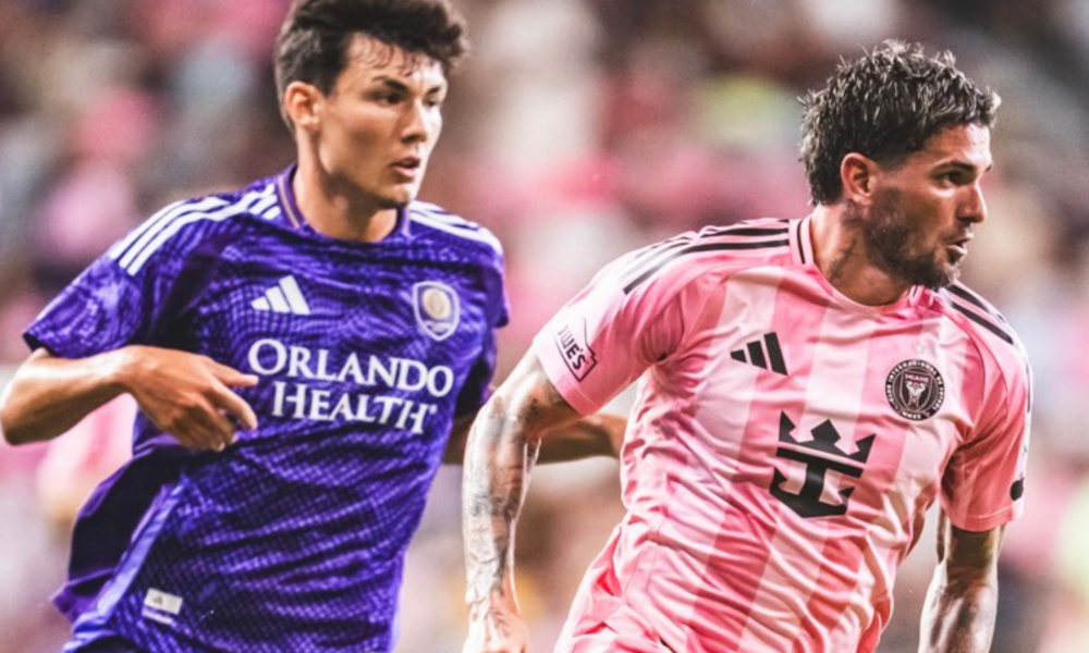 Two soccer players chase the ball on the field, one in a purple Orlando Health jersey and the other in a pink striped kit.
