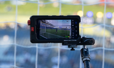 Close-up of a rugged monitor mounted on a tripod filming a soccer goal in a stadium, net visible on screen.