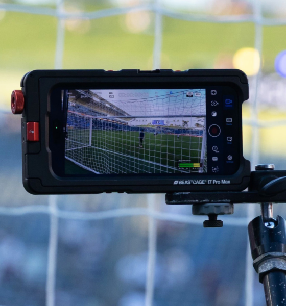 Close-up of a rugged monitor mounted on a tripod filming a soccer goal in a stadium, net visible on screen.