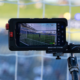 Close-up of a rugged monitor mounted on a tripod filming a soccer goal in a stadium, net visible on screen.