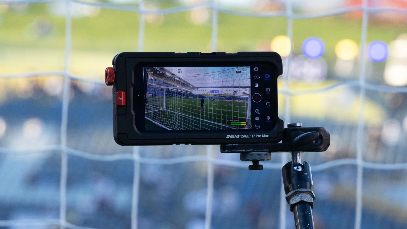 Close-up of a rugged monitor mounted on a tripod filming a soccer goal in a stadium, net visible on screen.