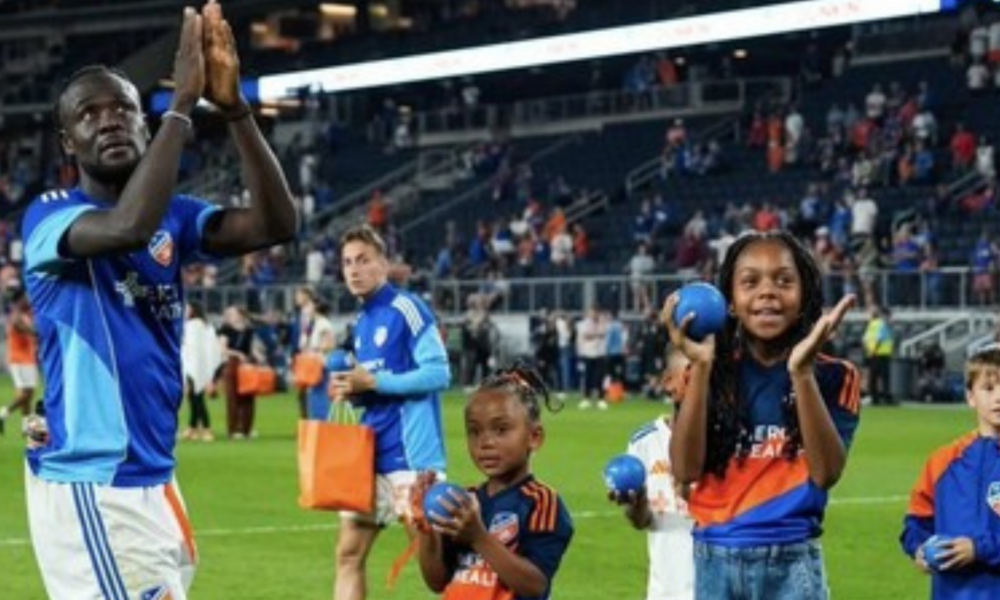 Soccer players and children on a green field after a match; a tall blue-clad player claps as kids hold balls and gift bags with the stands in the background.