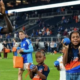 Soccer players and children on a green field after a match; a tall blue-clad player claps as kids hold balls and gift bags with the stands in the background.