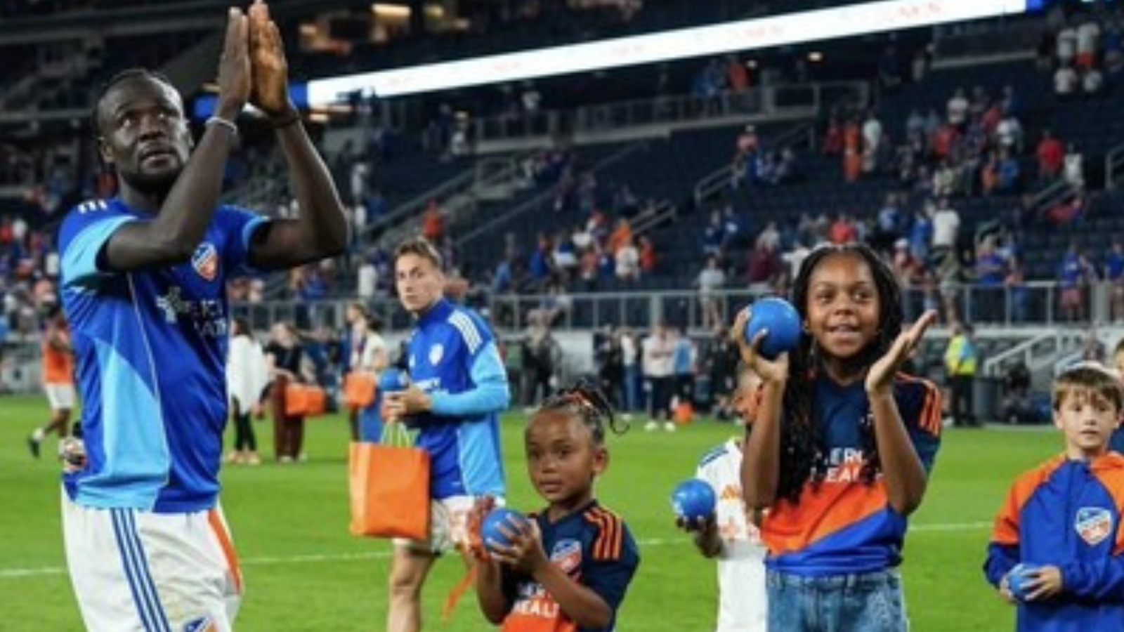 Soccer players and children on a green field after a match; a tall blue-clad player claps as kids hold balls and gift bags with the stands in the background.