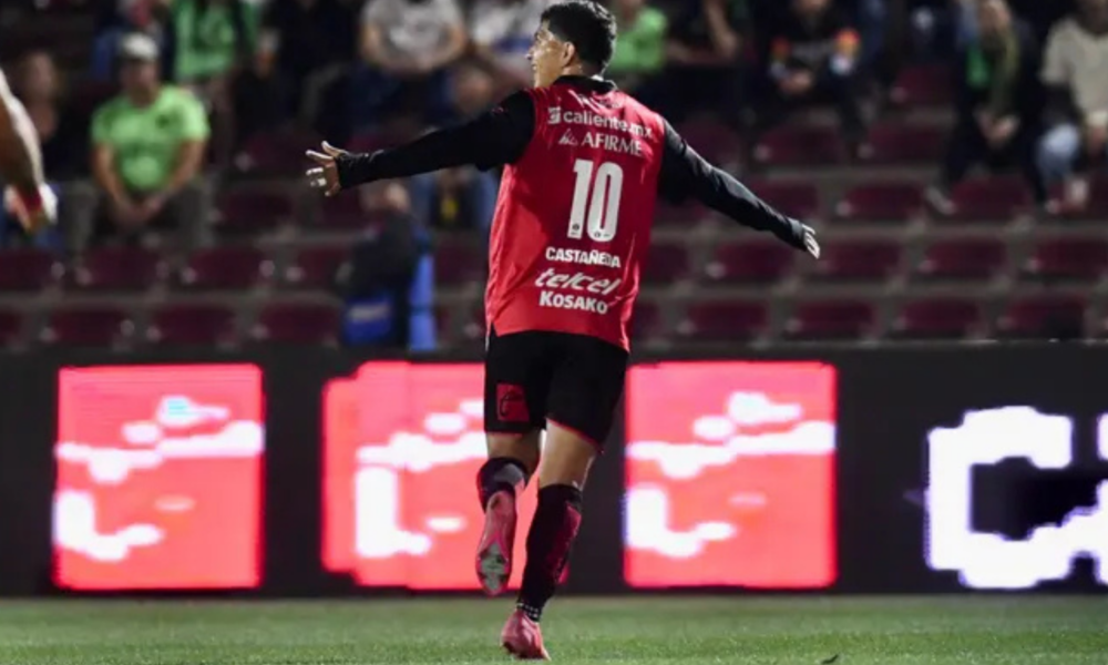 Soccer player in a red jersey with the number 10, arms outstretched, celebrating on the field with stadium seats and LED boards in the background.