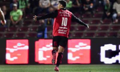Soccer player in a red jersey with the number 10, arms outstretched, celebrating on the field with stadium seats and LED boards in the background.