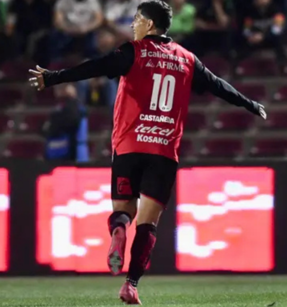 Soccer player in a red jersey with the number 10, arms outstretched, celebrating on the field with stadium seats and LED boards in the background.