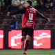 Soccer player in a red jersey with the number 10, arms outstretched, celebrating on the field with stadium seats and LED boards in the background.