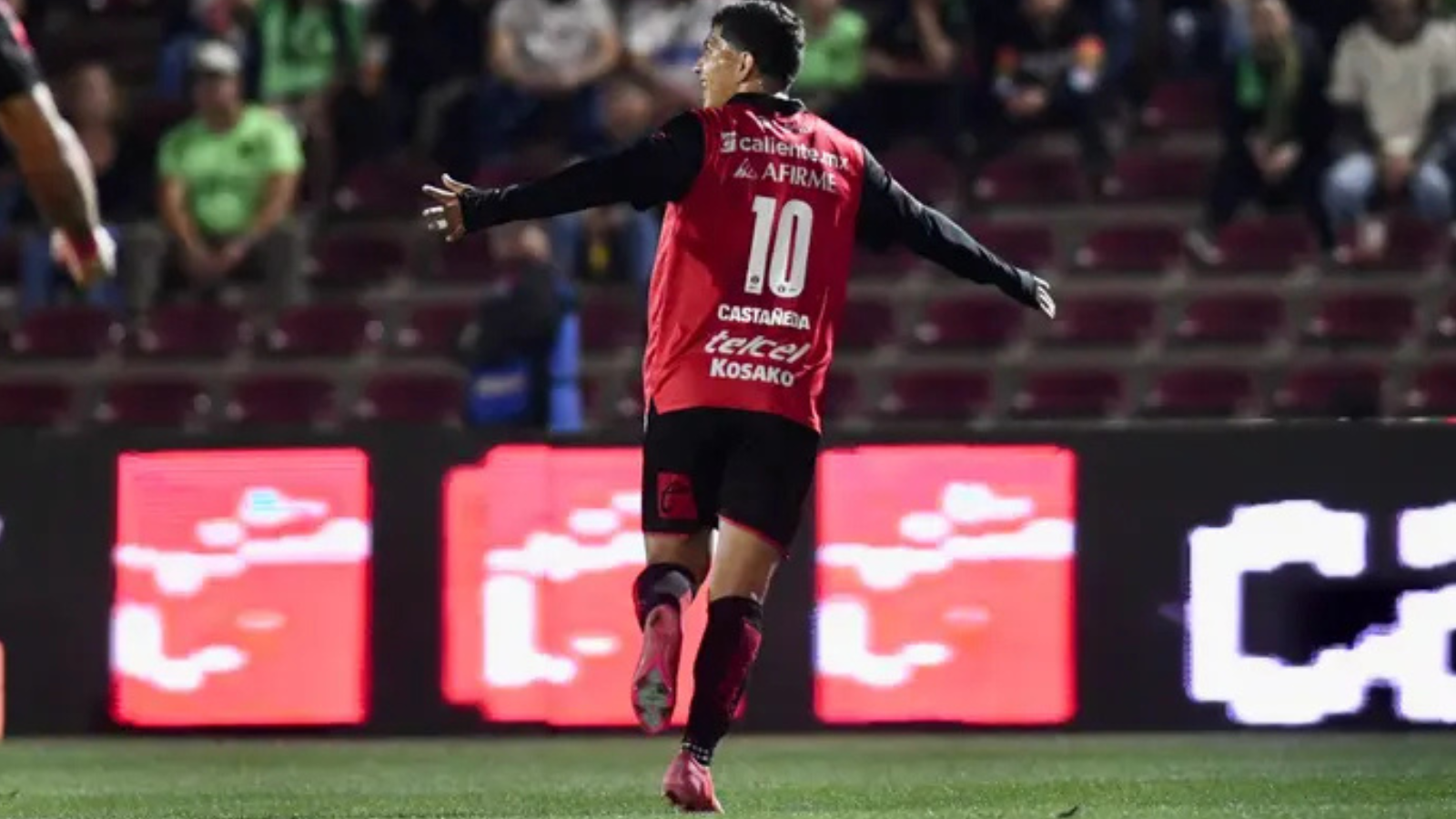 Soccer player in a red jersey with the number 10, arms outstretched, celebrating on the field with stadium seats and LED boards in the background.