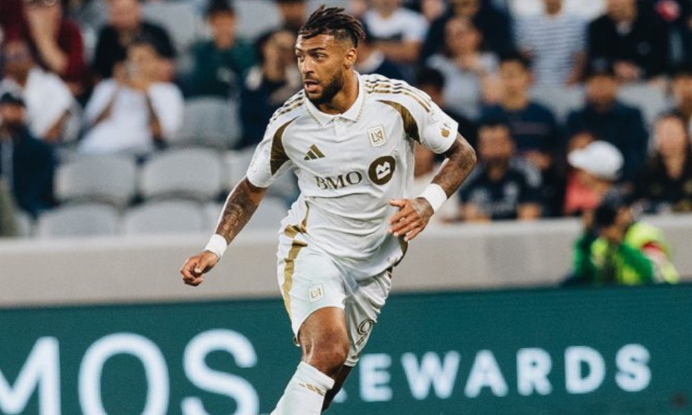 Soccer player in a white LAFC jersey sprinting on the pitch with a cheering crowd in the background.