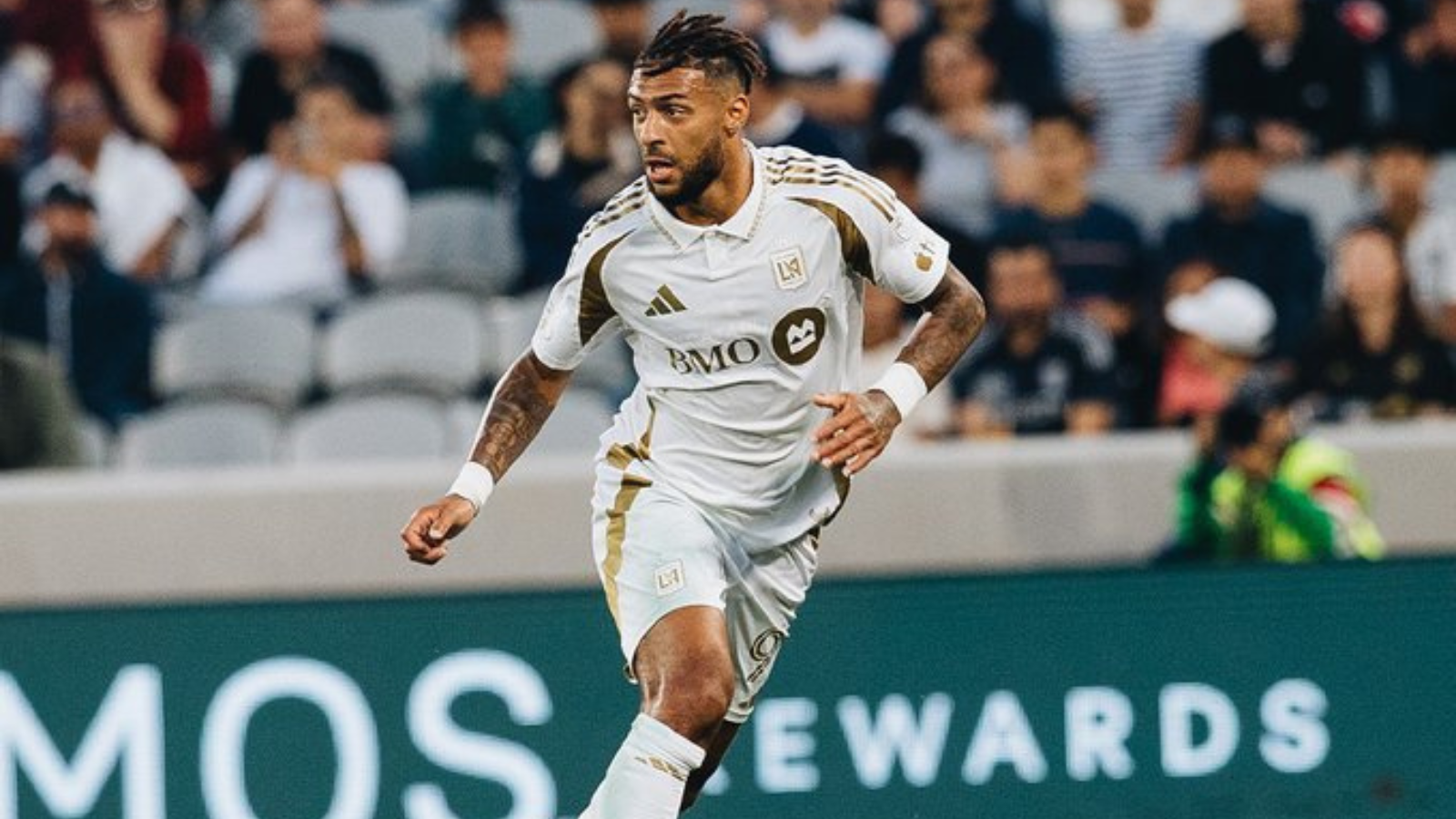 Soccer player in a white LAFC jersey sprinting on the pitch with a cheering crowd in the background.