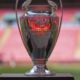 Shiny silver trophy on a black pedestal in a stadium, with blurred pink seating in the background.