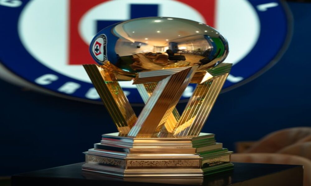 Golden trophy with interlocking angular supports and a reflective spherical top on a tiered pedestal, placed on a dark table in a room with a blurred logo in the background.