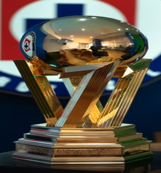 Golden trophy with interlocking angular supports and a reflective spherical top on a tiered pedestal, placed on a dark table in a room with a blurred logo in the background.