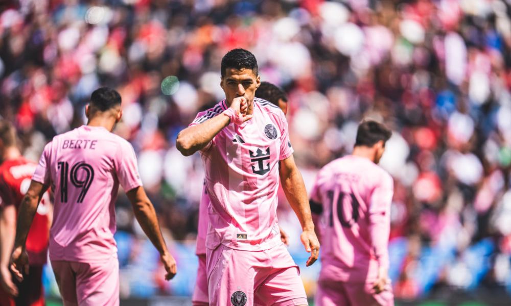Soccer player in pink kit celebrating, kissing his hand, with teammates and a packed stadium behind.