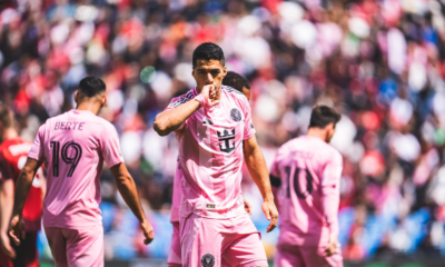 Soccer player in pink kit celebrating, kissing his hand, with teammates and a packed stadium behind.