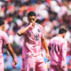 Soccer player in pink kit celebrating, kissing his hand, with teammates and a packed stadium behind.