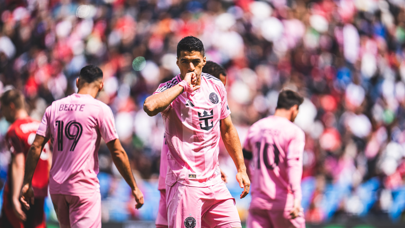 Soccer player in pink kit celebrating, kissing his hand, with teammates and a packed stadium behind.