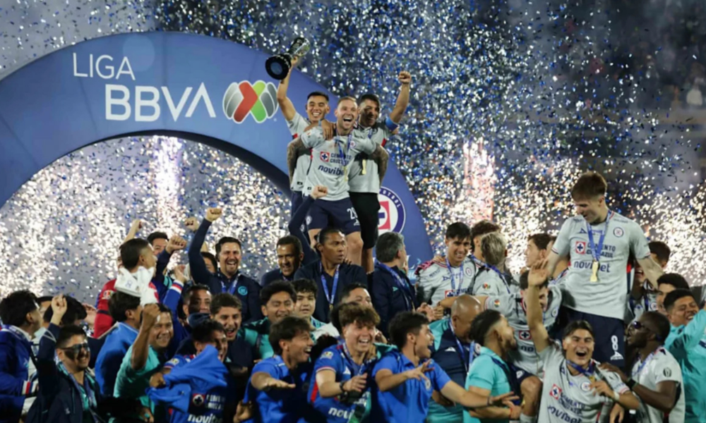 Three players on a podium lift a trophy while teammates cheer beneath a blue LIGA BBVA arch as confetti rains down in celebration of a league title.