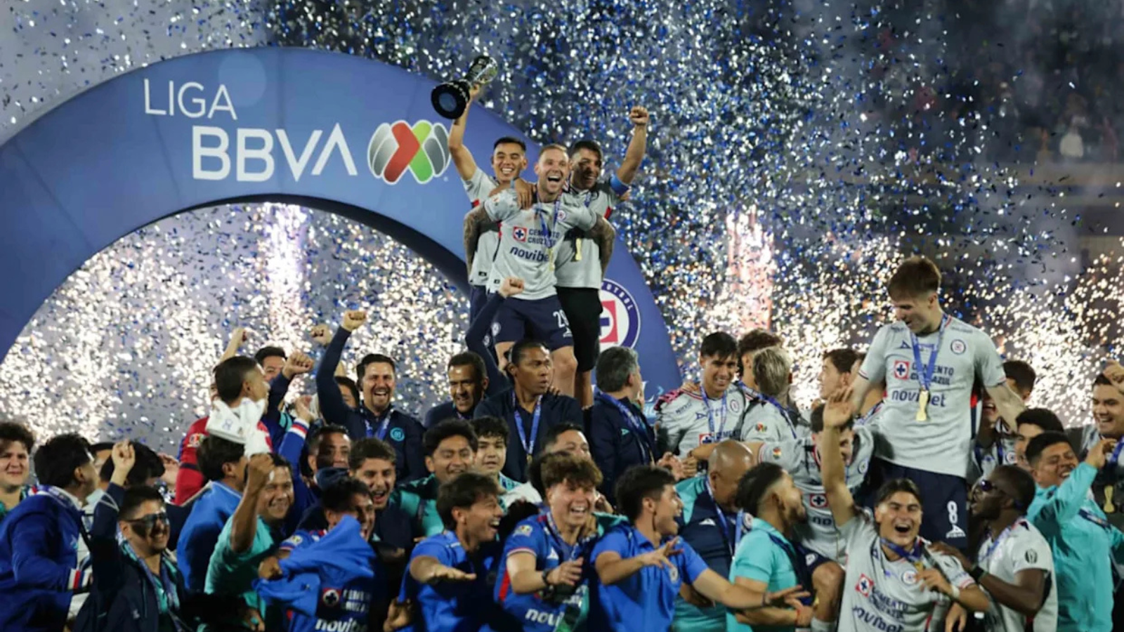 Three players on a podium lift a trophy while teammates cheer beneath a blue LIGA BBVA arch as confetti rains down in celebration of a league title.