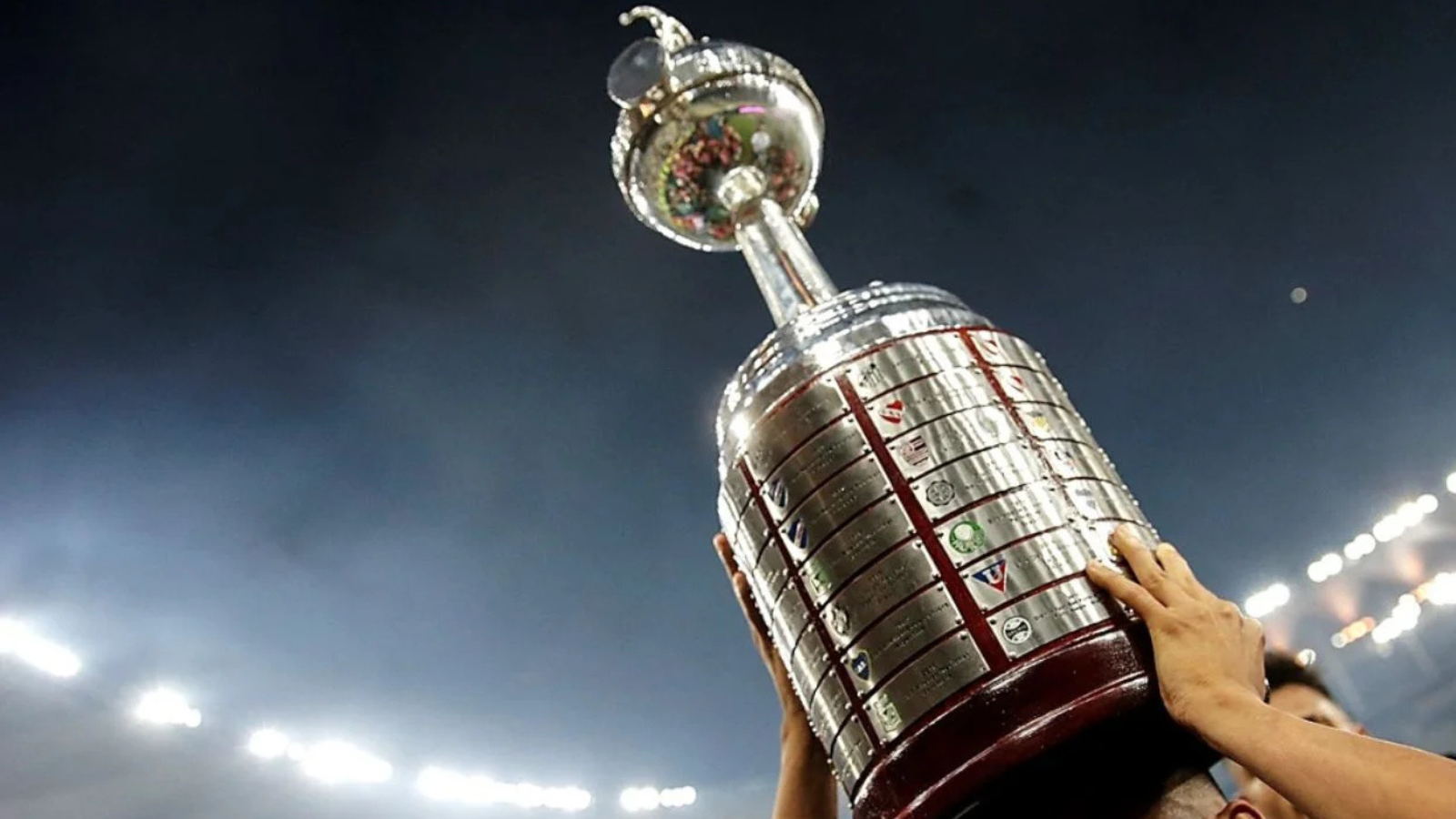 Athletes raise a tall, shiny silver championship trophy above their heads at night stadium celebration.
