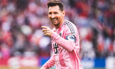 Soccer player in a pink striped jersey smiles and points during a match, with a blurred crowd in the background.