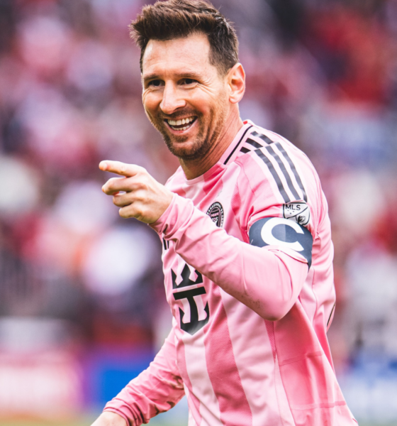 Soccer player in a pink striped jersey smiles and points during a match, with a blurred crowd in the background.