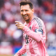 Soccer player in a pink striped jersey smiles and points during a match, with a blurred crowd in the background.