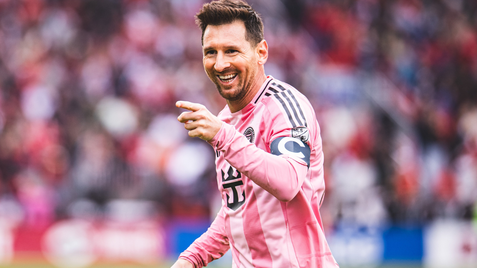 Soccer player in a pink striped jersey smiles and points during a match, with a blurred crowd in the background.