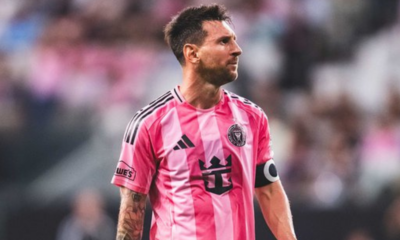 Soccer player in a pink striped jersey with a captain's armband looking to the right on the field.