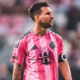 Soccer player in a pink striped jersey with a captain's armband looking to the right on the field.