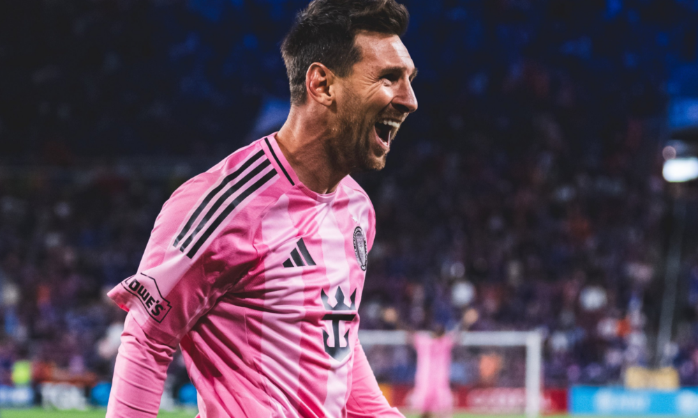Male soccer player in a pink jersey celebrating with mouth open on a field, crowd in the background