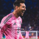 Male soccer player in a pink jersey celebrating with mouth open on a field, crowd in the background