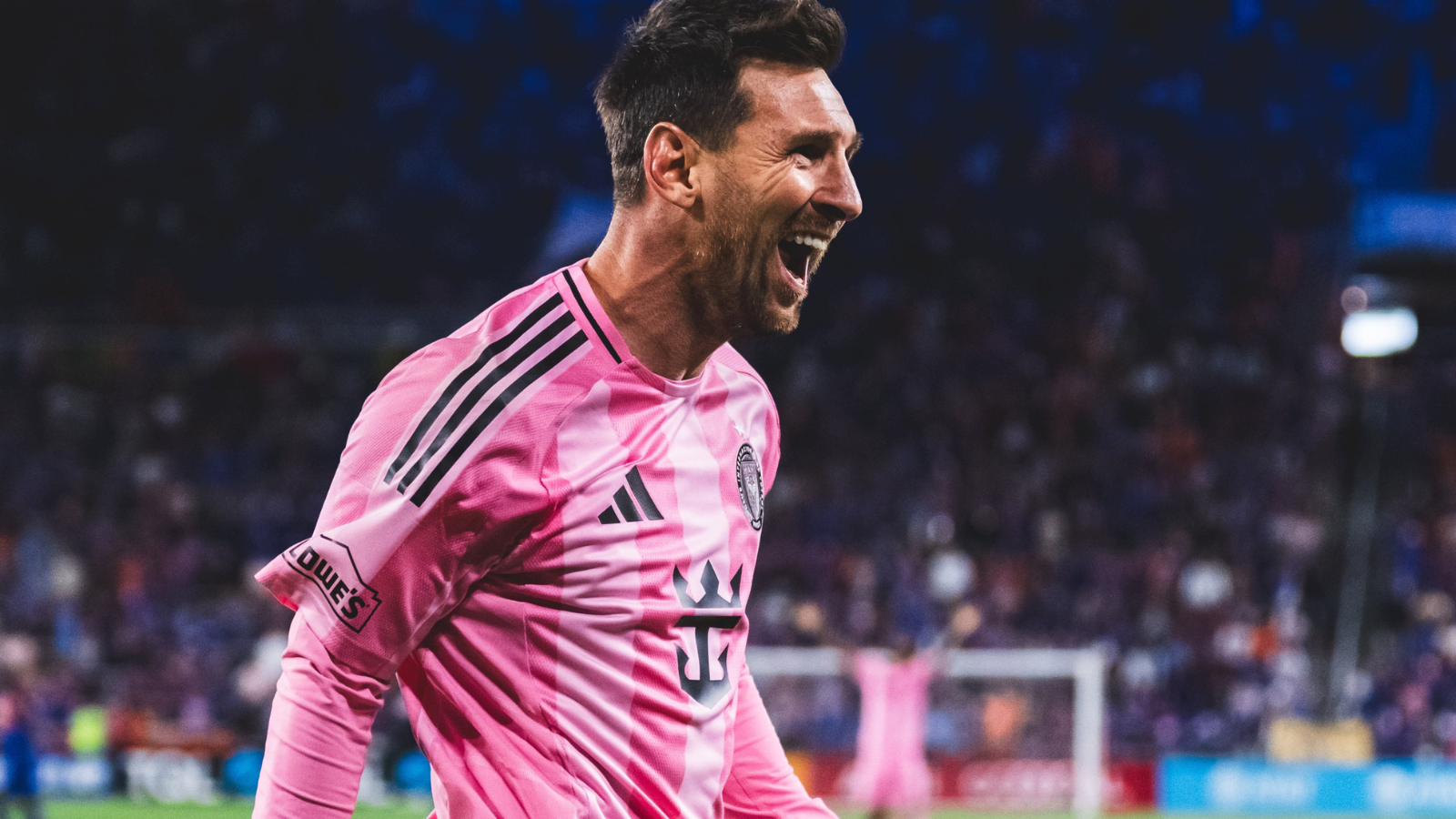 Male soccer player in a pink jersey celebrating with mouth open on a field, crowd in the background