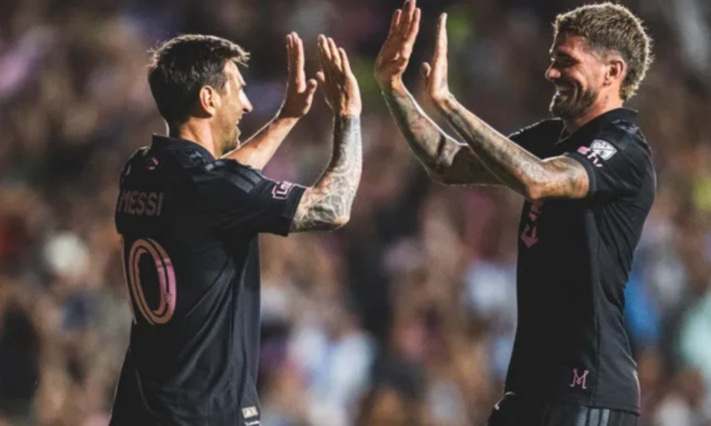 Two soccer players in black jerseys high-five each other on the field, with a cheering crowd in the background (one wearing number 10 on the back).