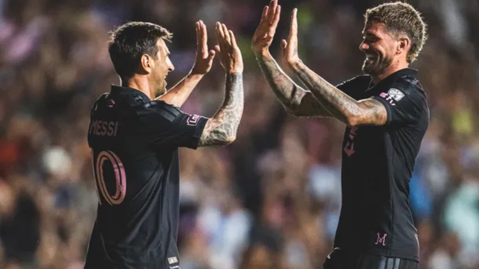 Two soccer players in black jerseys high-five each other on the field, with a cheering crowd in the background (one wearing number 10 on the back).