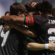 Soccer players in black jerseys hug in a group celebration on the field.