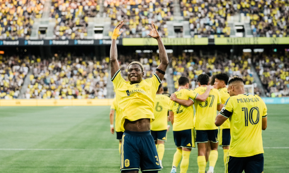 Soccer players in yellow jerseys celebrate a goal on the field as fans cheer in the stands, with one player raising his arms in victory.