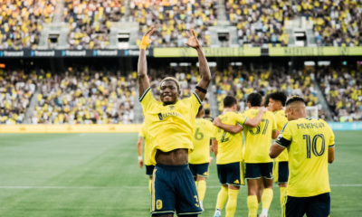 Soccer players in yellow jerseys celebrate a goal on the field as fans cheer in the stands, with one player raising his arms in victory.